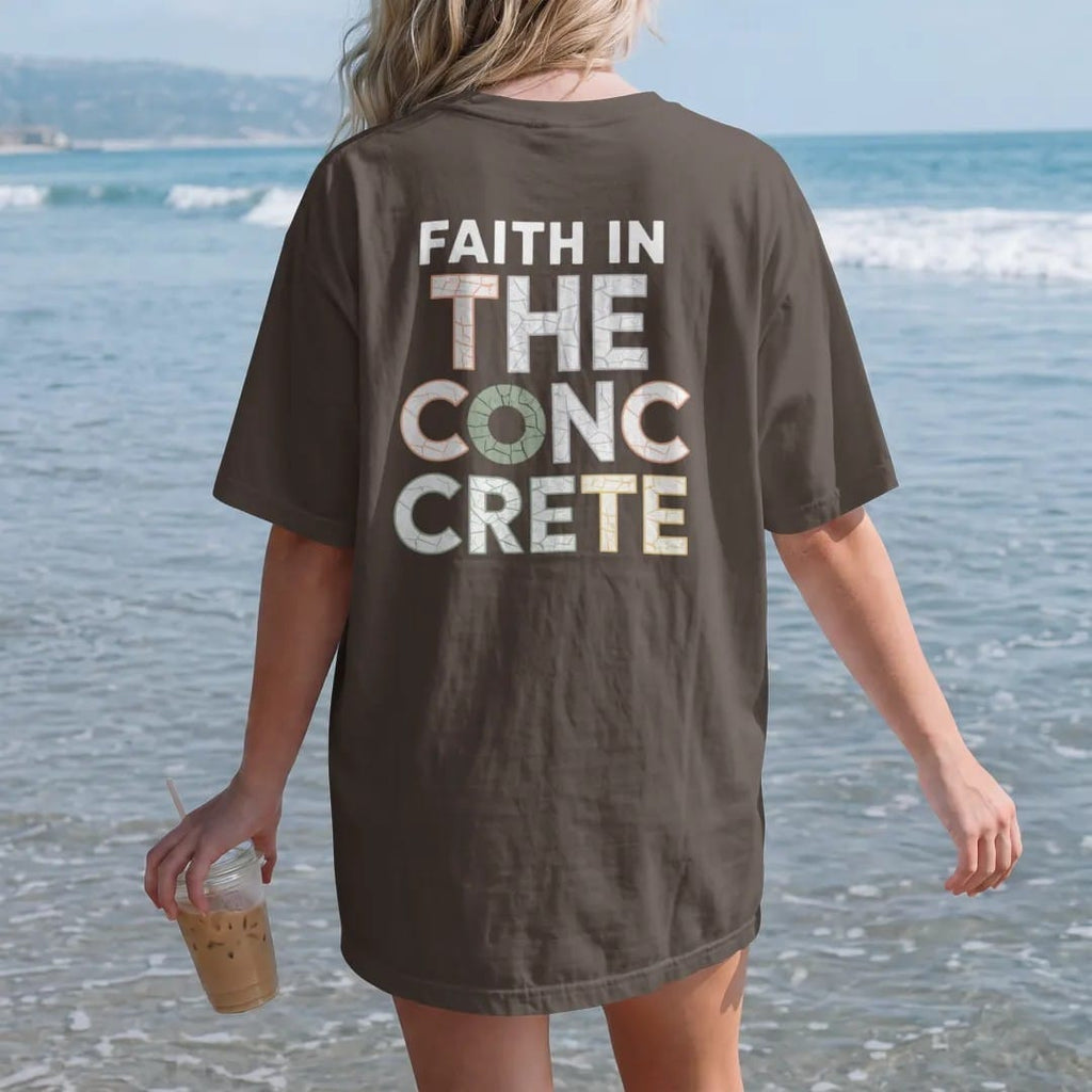 Woman on the beach in a Espresso tee, back view: large cracked-concrete letters reading The Concrete under small spaced Faith In with a halo over the first letter, in bone white, orange and green.