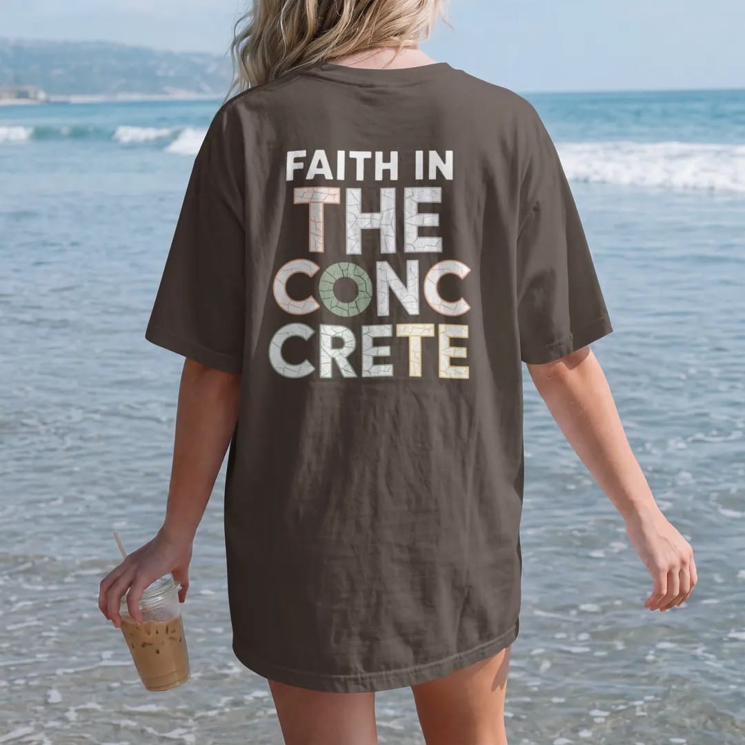 Woman on the beach in a Espresso tee, back view: large cracked-concrete letters reading The Concrete under small spaced Faith In with a halo over the first letter, in bone white, orange and green.