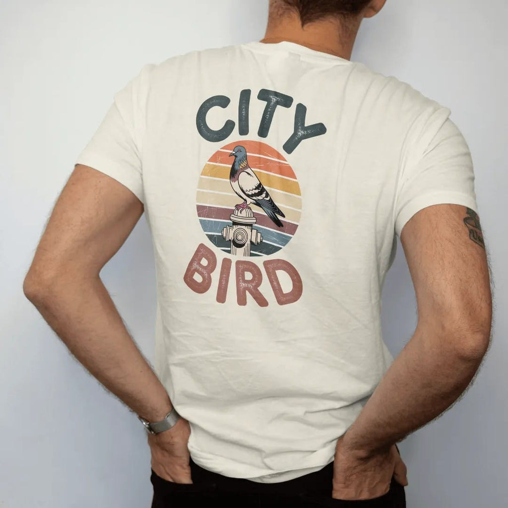 Man wearing a Ivory tee, seen from the back: a city pigeon perched on a fire hydrant in front of a faded retro sunset with rainbow stripes, between chunky 70s letters reading City and Bird, with a worn, cracked-ink vintage finish.