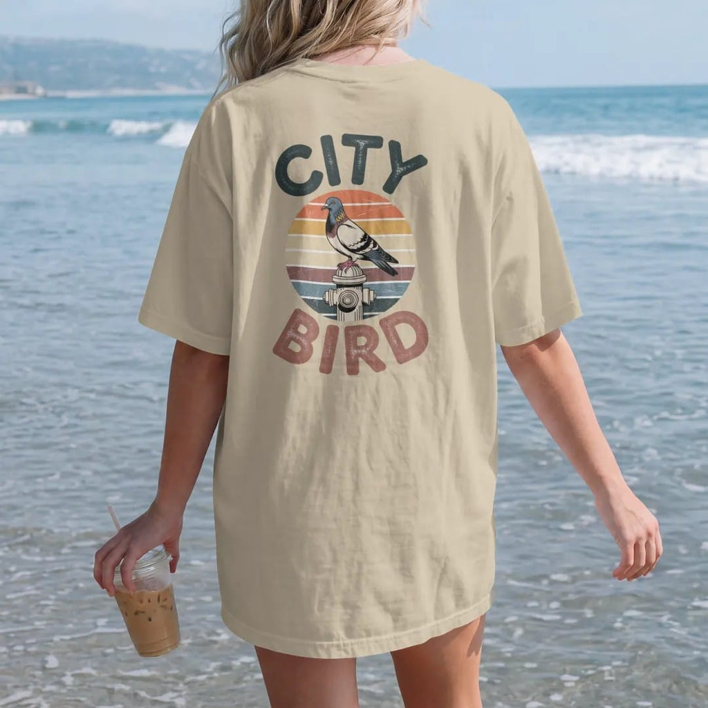 Woman on the beach in a Sandstone tee, back view: a city pigeon perched on a fire hydrant in front of a faded retro sunset with rainbow stripes, between chunky 70s letters reading City and Bird, with a worn, cracked-ink vintage finish.