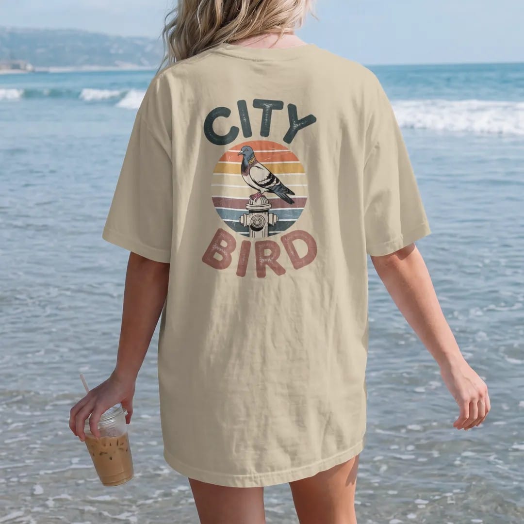 Woman on the beach in a Sandstone tee, back view: a city pigeon perched on a fire hydrant in front of a faded retro sunset with rainbow stripes, between chunky 70s letters reading City and Bird, with a worn, cracked-ink vintage finish.
