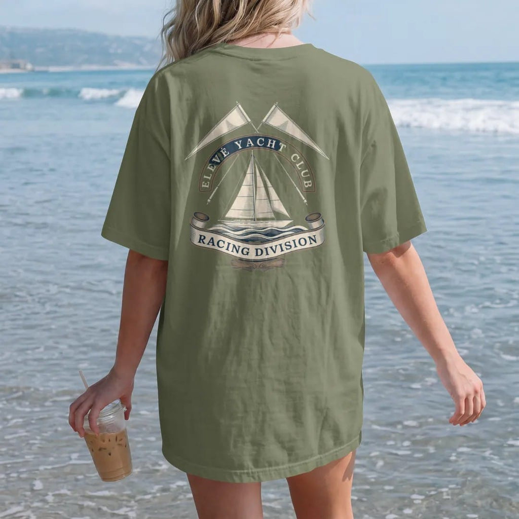 Woman on the beach in a Moss tee, back view: a sailing yacht on stylized waves under crossed pennants, with Elevé Yacht Club arched above and Racing Division below, as a fine engraving in cream and champagne gold.