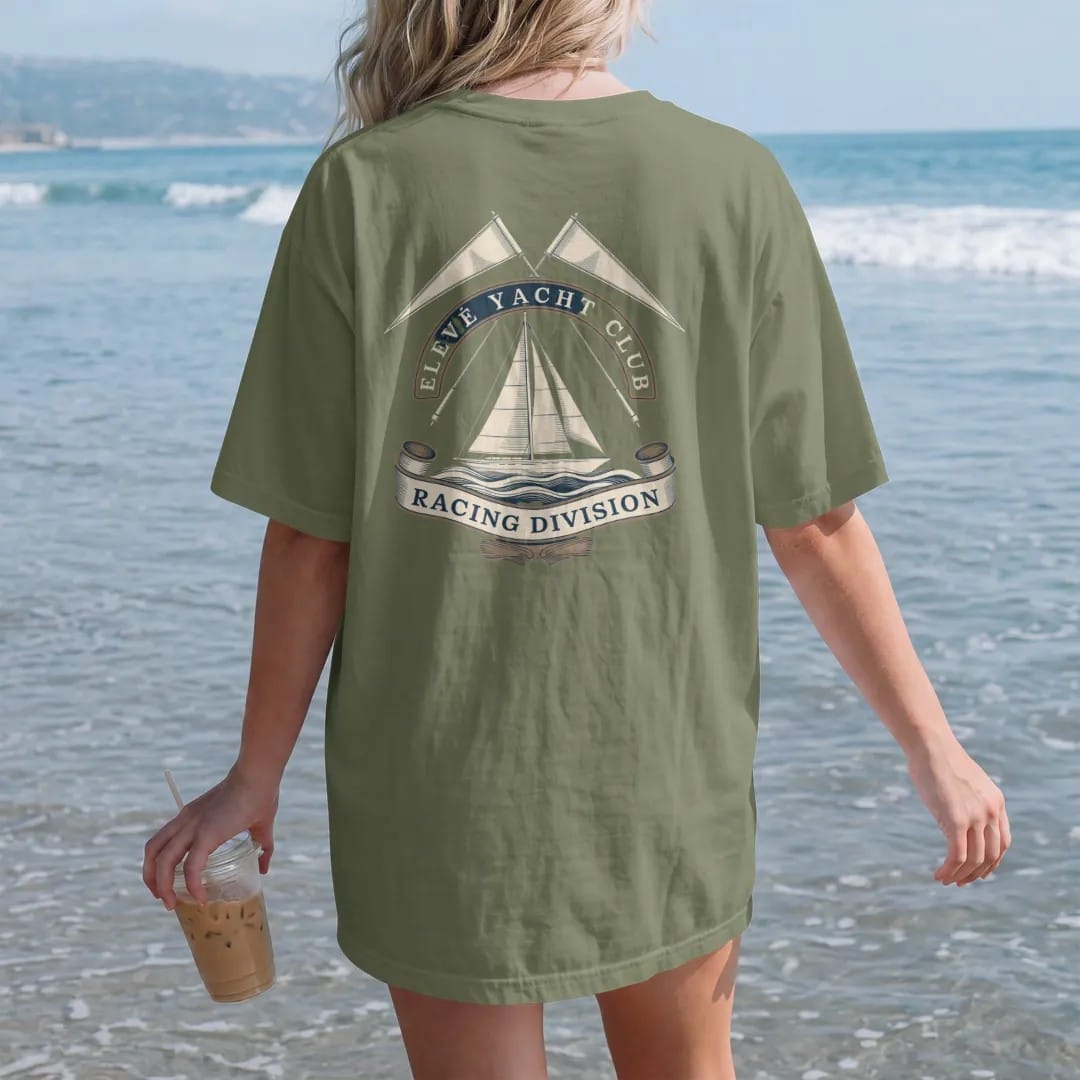 Woman on the beach in a Moss tee, back view: a sailing yacht on stylized waves under crossed pennants, with Elevé Yacht Club arched above and Racing Division below, as a fine engraving in cream and champagne gold.