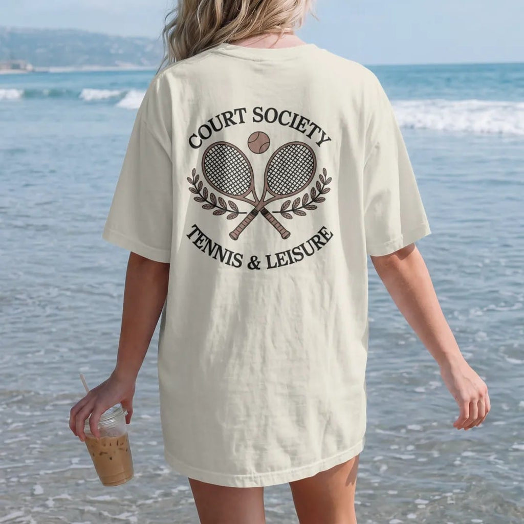 Woman on the beach in a Ivory tee, back view: two crossed wooden tennis rackets with a ball and laurel sprigs under Court Society and Tennis & Leisure, as a fine engraving in navy and antique gold.