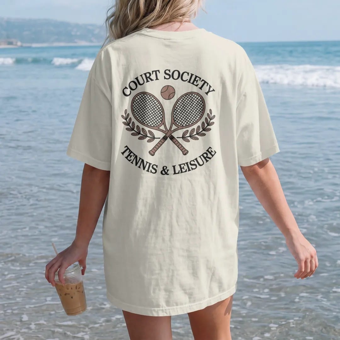Woman on the beach in a Ivory tee, back view: two crossed wooden tennis rackets with a ball and laurel sprigs under Court Society and Tennis & Leisure, as a fine engraving in navy and antique gold.