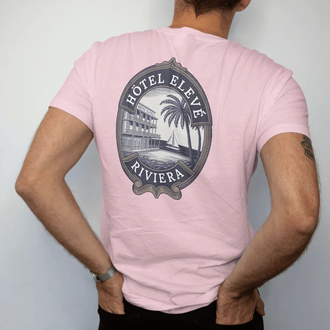 Man wearing a Blossom tee, seen from the back: an oval vintage luggage label with a seaside hotel, palms and a sailboat, titled Hôtel Elevé with a Riviera banner, as a fine engraving in navy and antique gold.