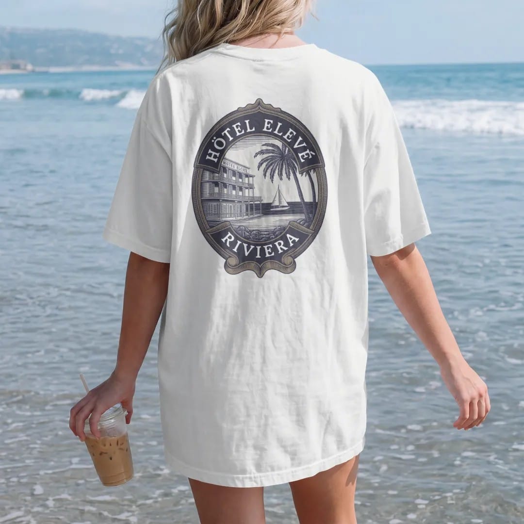 Woman on the beach in a White tee, back view: an oval vintage luggage label with a seaside hotel, palms and a sailboat, titled Hôtel Elevé with a Riviera banner, as a fine engraving in navy and antique gold.