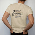 Man wearing a Sandstone tee, seen from the back: a minimal wordmark with italic Quietly above spaced Exceptional, a thin rule and a tiny laurel sprig, as a fine engraving in navy and antique gold.