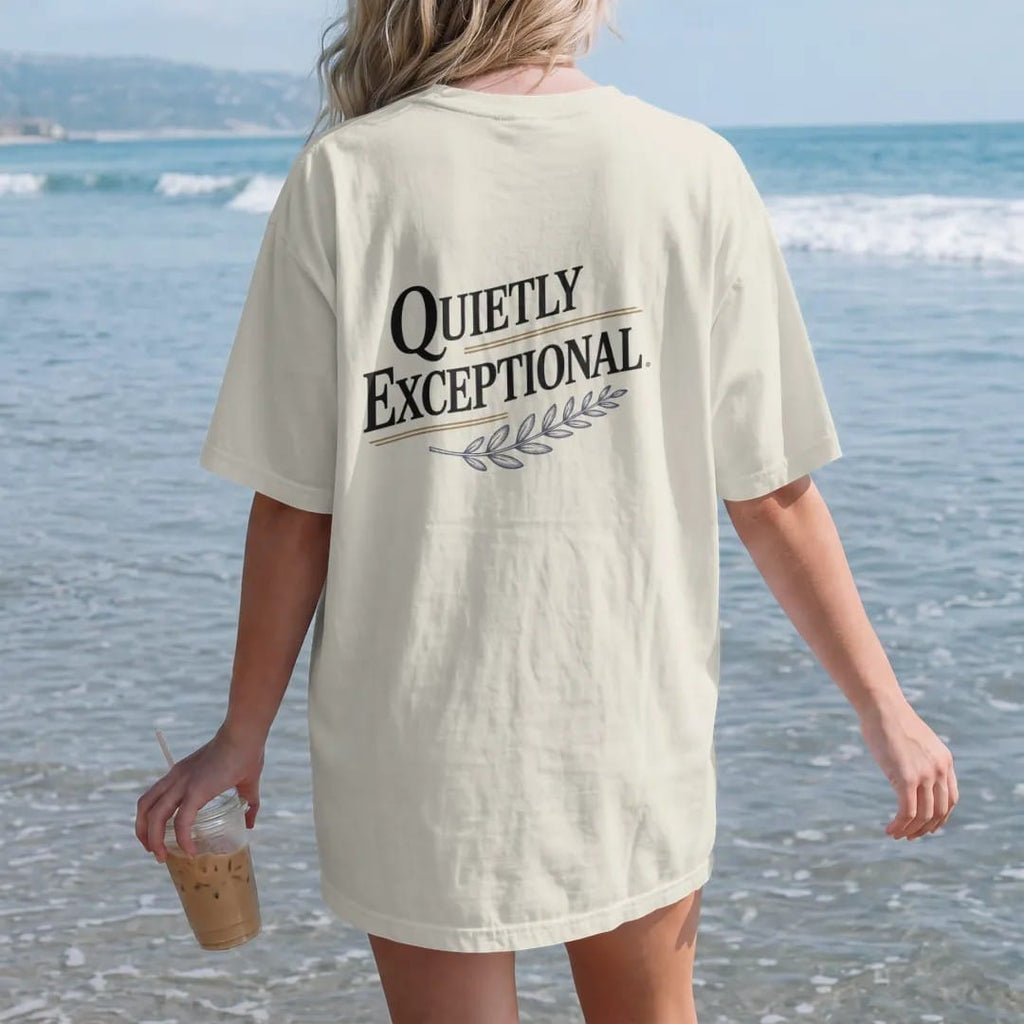 Woman on the beach in a Ivory tee, back view: a minimal wordmark with italic Quietly above spaced Exceptional, a thin rule and a tiny laurel sprig, as a fine engraving in navy and antique gold.