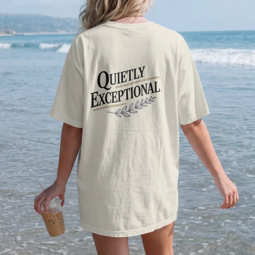 Woman on the beach in a Ivory tee, back view: a minimal wordmark with italic Quietly above spaced Exceptional, a thin rule and a tiny laurel sprig, as a fine engraving in navy and antique gold.