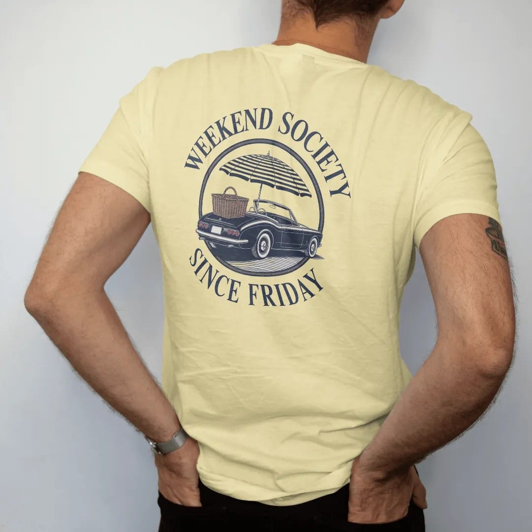 Man wearing a Butter tee, seen from the back: a vintage convertible with a picnic basket and umbrella in a round emblem, ringed by Weekend Society and Since Friday, as a fine engraving in navy and antique gold.