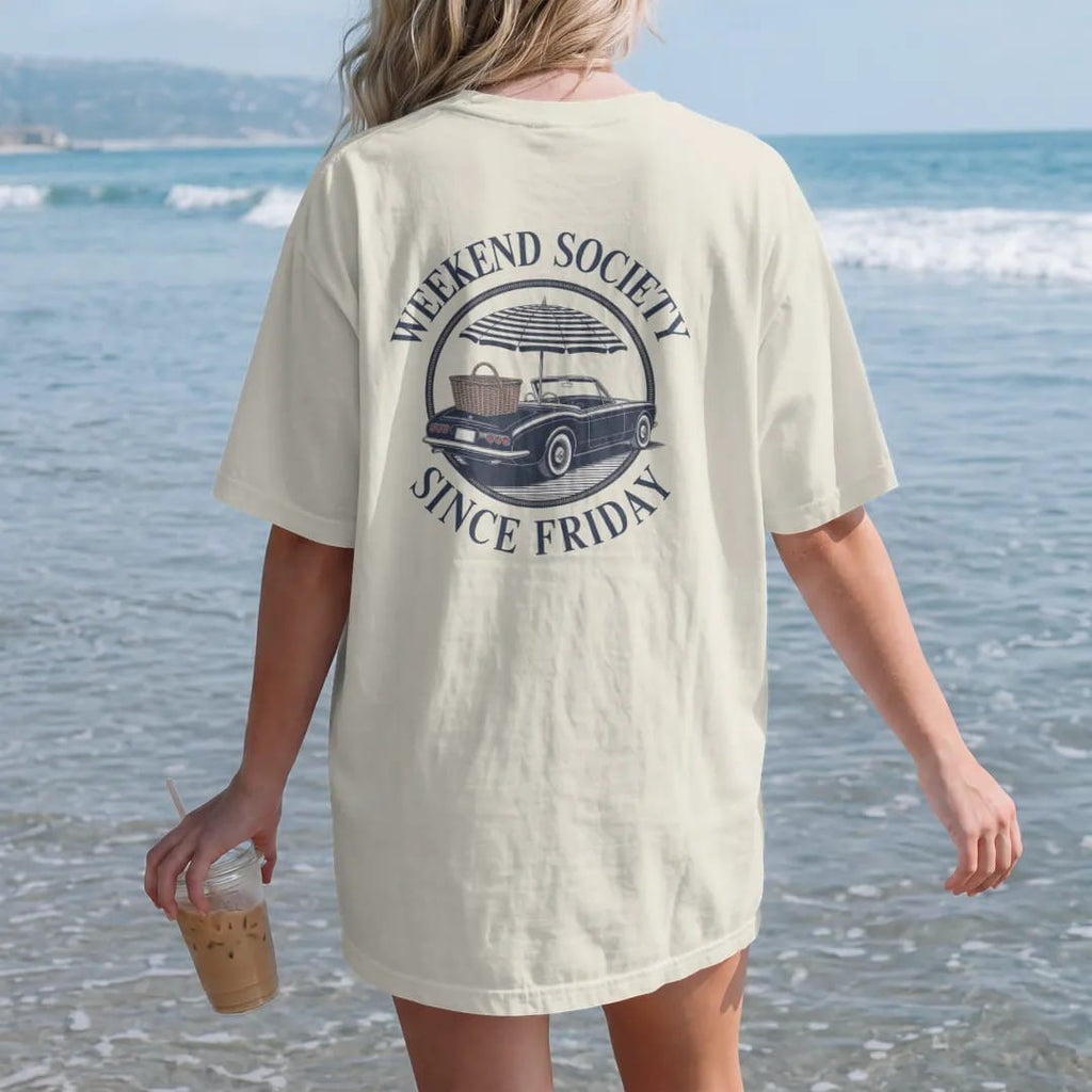Woman on the beach in a Ivory tee, back view: a vintage convertible with a picnic basket and umbrella in a round emblem, ringed by Weekend Society and Since Friday, as a fine engraving in navy and antique gold.