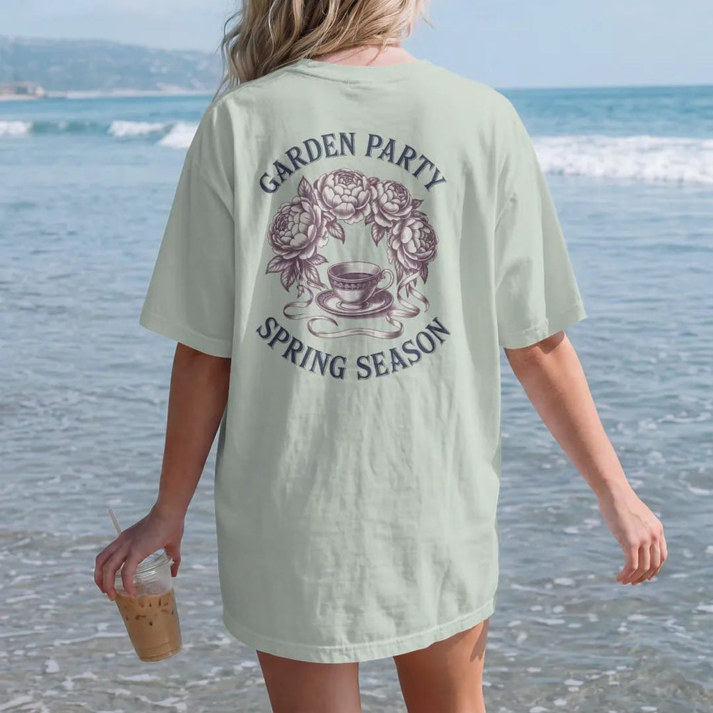Woman on the beach in a Bay tee, back view: a wreath of peonies and roses with ribbons around a teacup, with Garden Party in script and Spring Season below, as a fine engraving in navy and antique gold.
