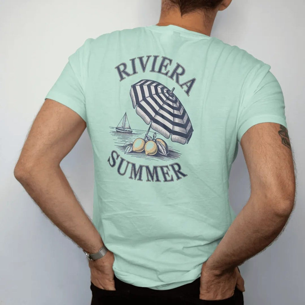 Man wearing a Chalky Mint tee, seen from the back: lemons on a branch, a striped beach umbrella and a sailboat on calm water under Riviera and Summer, as a fine engraving in navy and antique gold.