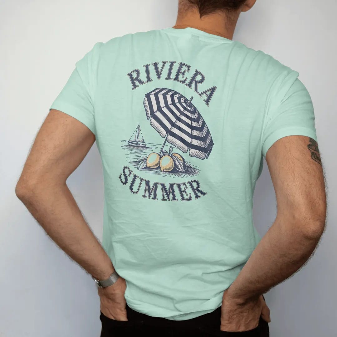 Man wearing a Chalky Mint tee, seen from the back: lemons on a branch, a striped beach umbrella and a sailboat on calm water under Riviera and Summer, as a fine engraving in navy and antique gold.