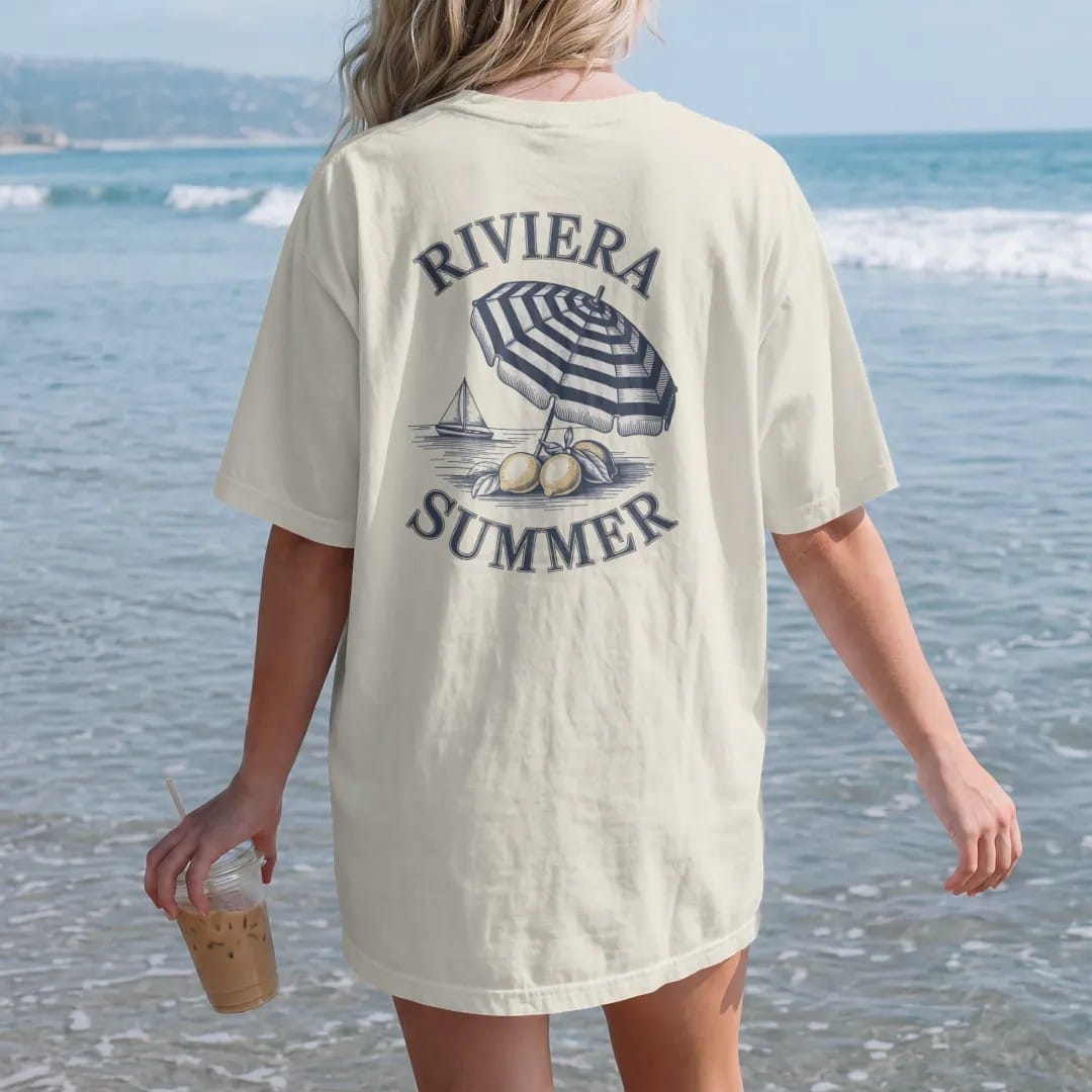 Woman on the beach in a Ivory tee, back view: lemons on a branch, a striped beach umbrella and a sailboat on calm water under Riviera and Summer, as a fine engraving in navy and antique gold.