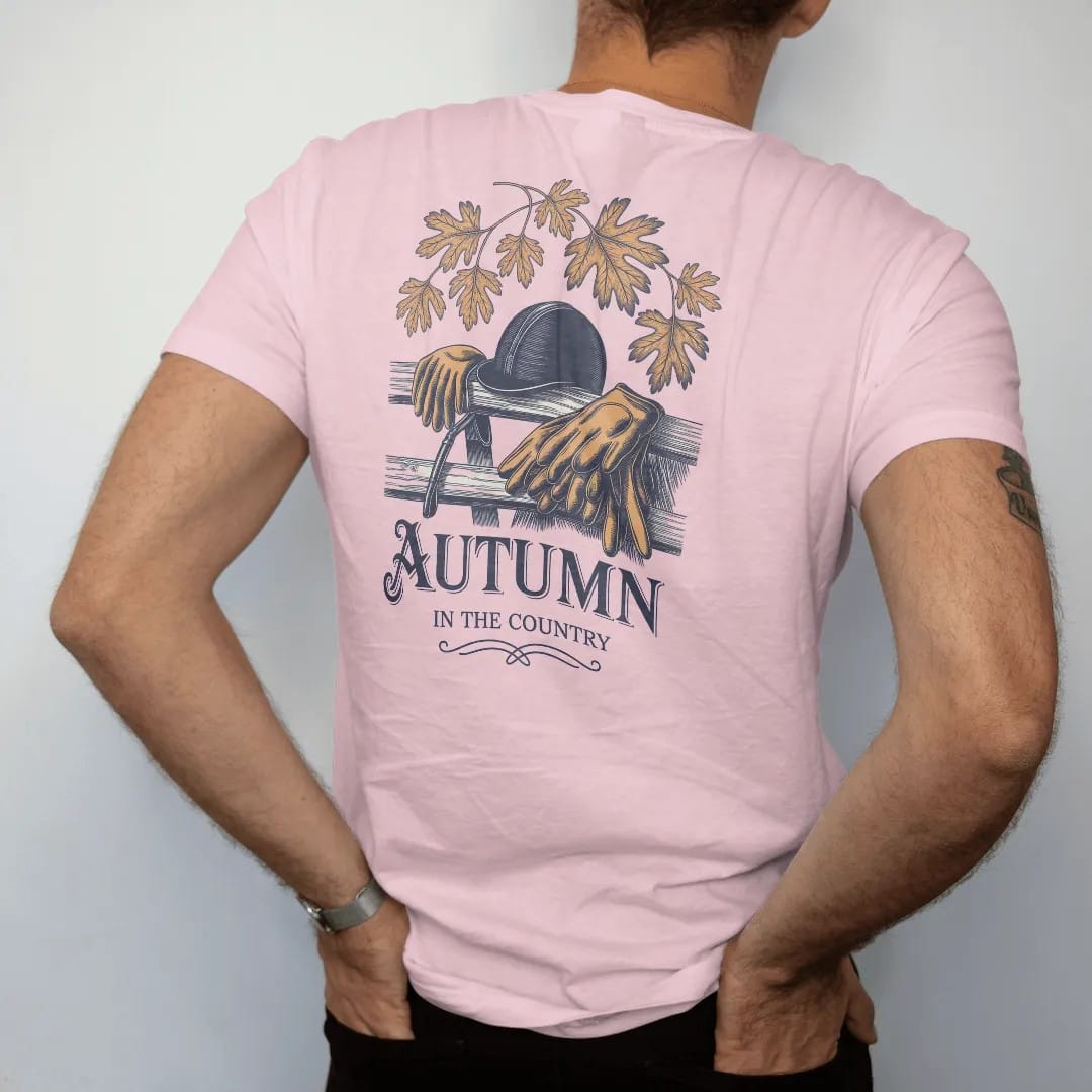 Man wearing a Blossom tee, seen from the back: a riding helmet, crop and gloves on a wooden fence with falling oak leaves, with Autumn in script and In The Country below, as a fine engraving in navy and antique gold.