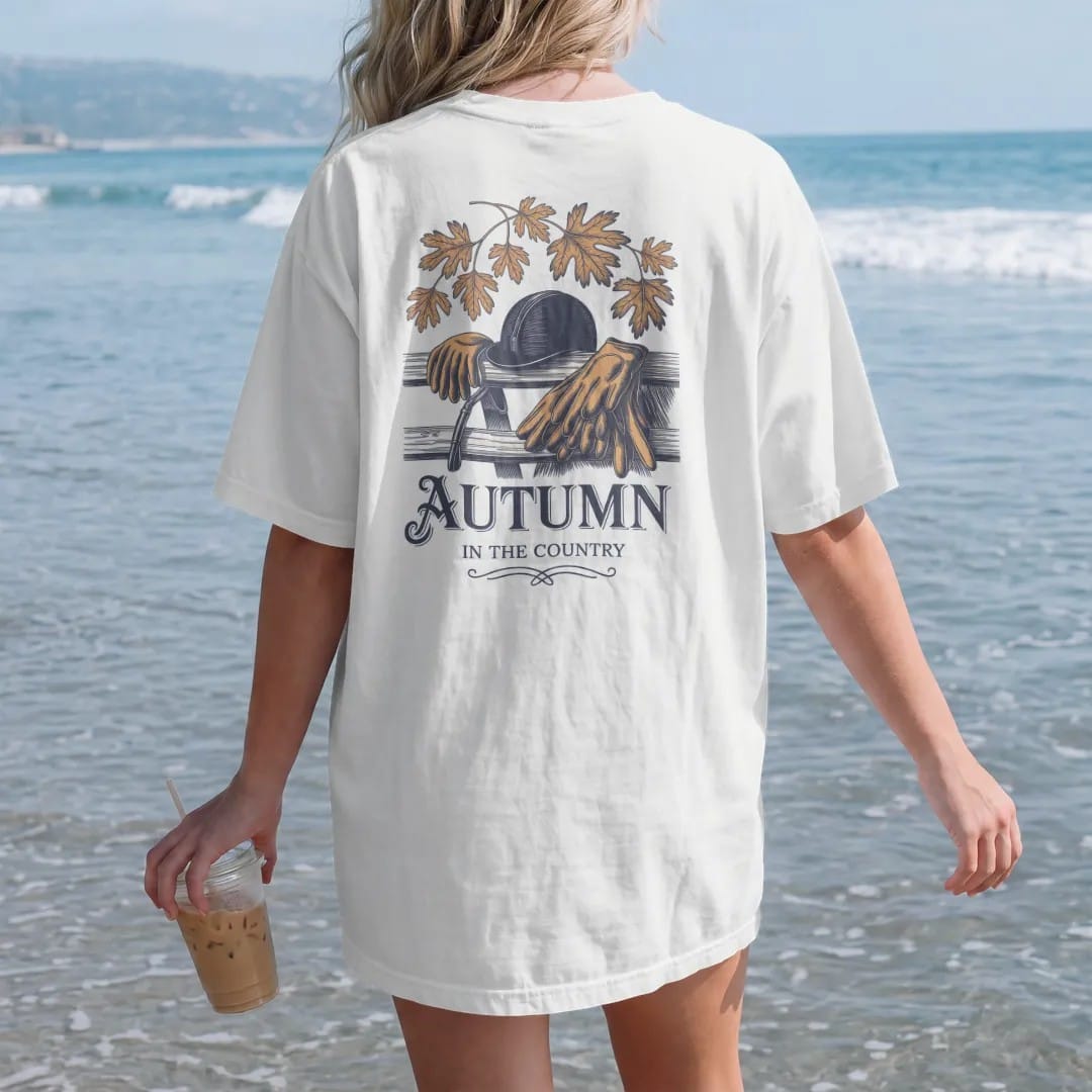 Woman on the beach in a White tee, back view: a riding helmet, crop and gloves on a wooden fence with falling oak leaves, with Autumn in script and In The Country below, as a fine engraving in navy and antique gold.