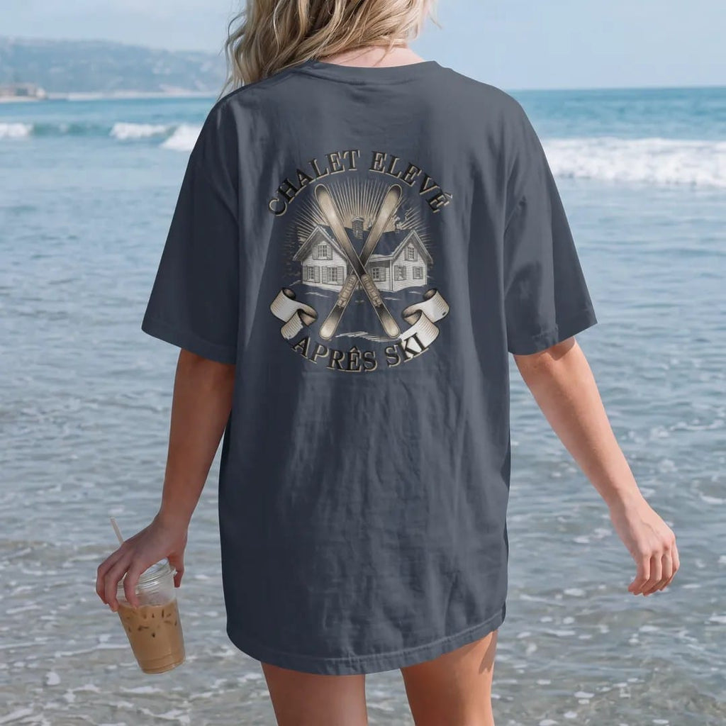 Woman on the beach in a Midnight tee, back view: a snowy alpine chalet with crossed vintage skis and pines under Chalet Elevé and an Après Ski banner, as a fine engraving in cream and champagne gold.