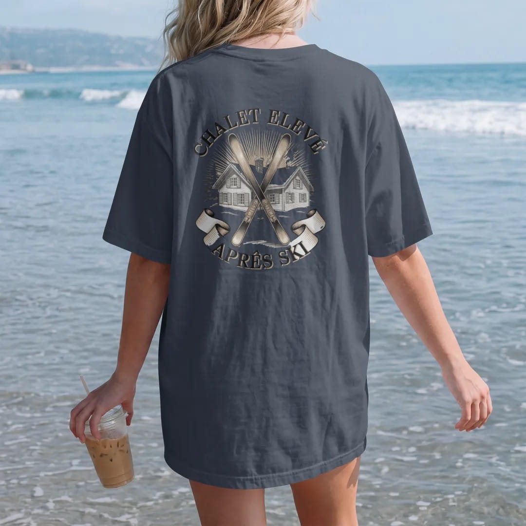 Woman on the beach in a Midnight tee, back view: a snowy alpine chalet with crossed vintage skis and pines under Chalet Elevé and an Après Ski banner, as a fine engraving in cream and champagne gold.