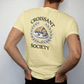 Man wearing a Butter tee, seen from the back: a golden croissant on a plate with a coffee cup inside a thin laurel, with Croissant arched above and Society below, as a fine engraving in navy and antique gold.