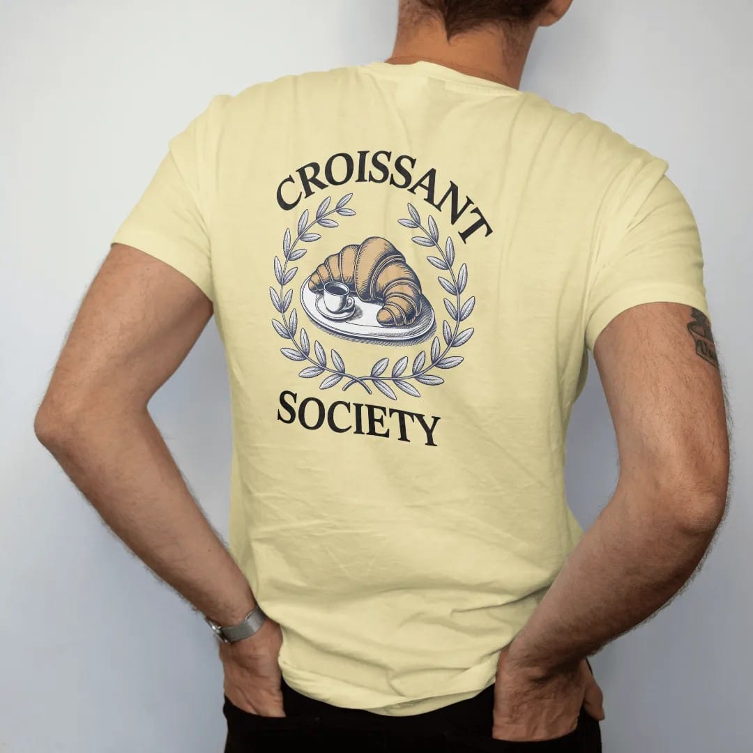 Man wearing a Butter tee, seen from the back: a golden croissant on a plate with a coffee cup inside a thin laurel, with Croissant arched above and Society below, as a fine engraving in navy and antique gold.