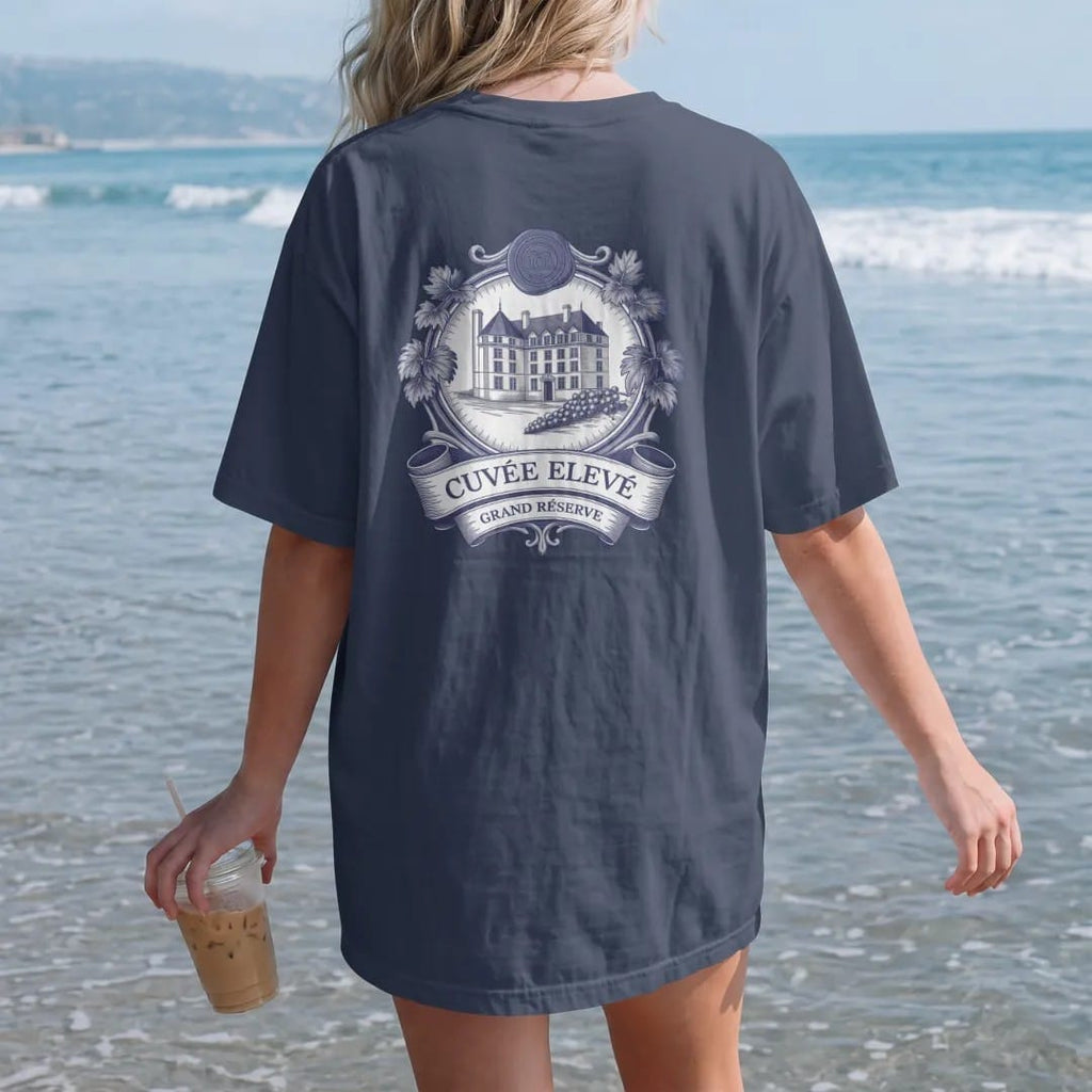 Woman on the beach in a Navy tee, back view: a vintage wine label with an engraved château, grapevines and a wax seal, reading Cuvée Elevé with a Grand Réserve banner, as a fine engraving in navy and antique gold.