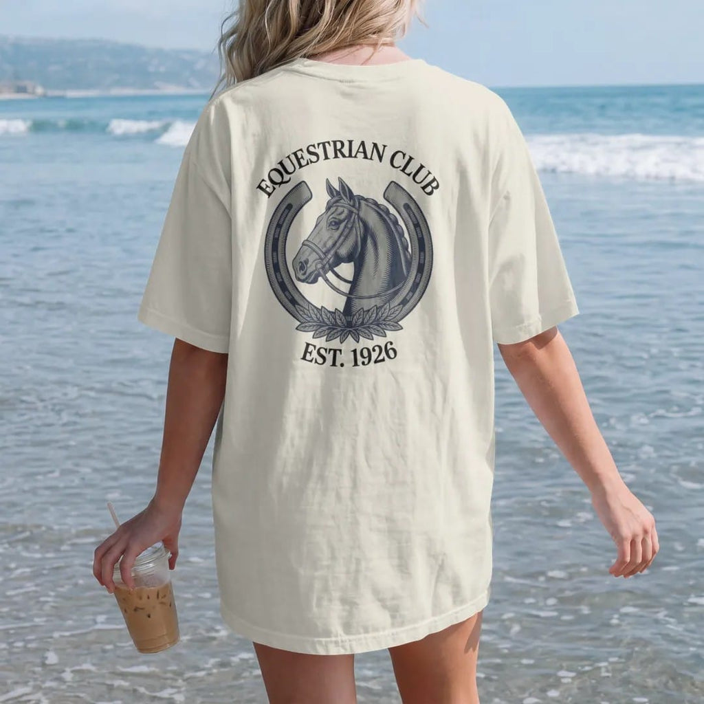 Woman on the beach in a Ivory tee, back view: a horse head in profile inside a horseshoe with a bridle and laurel, under Equestrian Club and Est. 1926, as a fine engraving in navy and antique gold.
