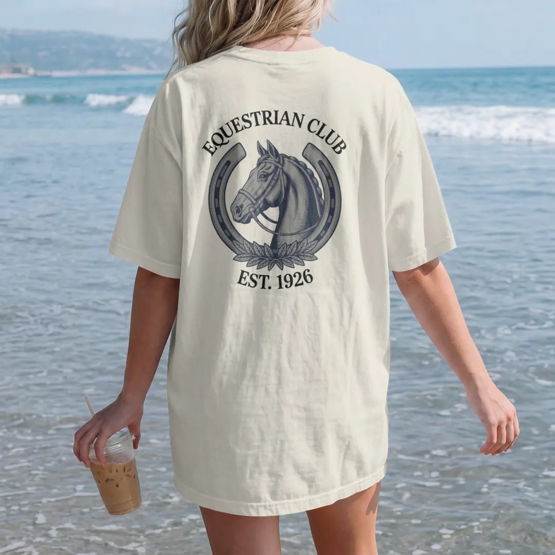 Woman on the beach in a Ivory tee, back view: a horse head in profile inside a horseshoe with a bridle and laurel, under Equestrian Club and Est. 1926, as a fine engraving in navy and antique gold.