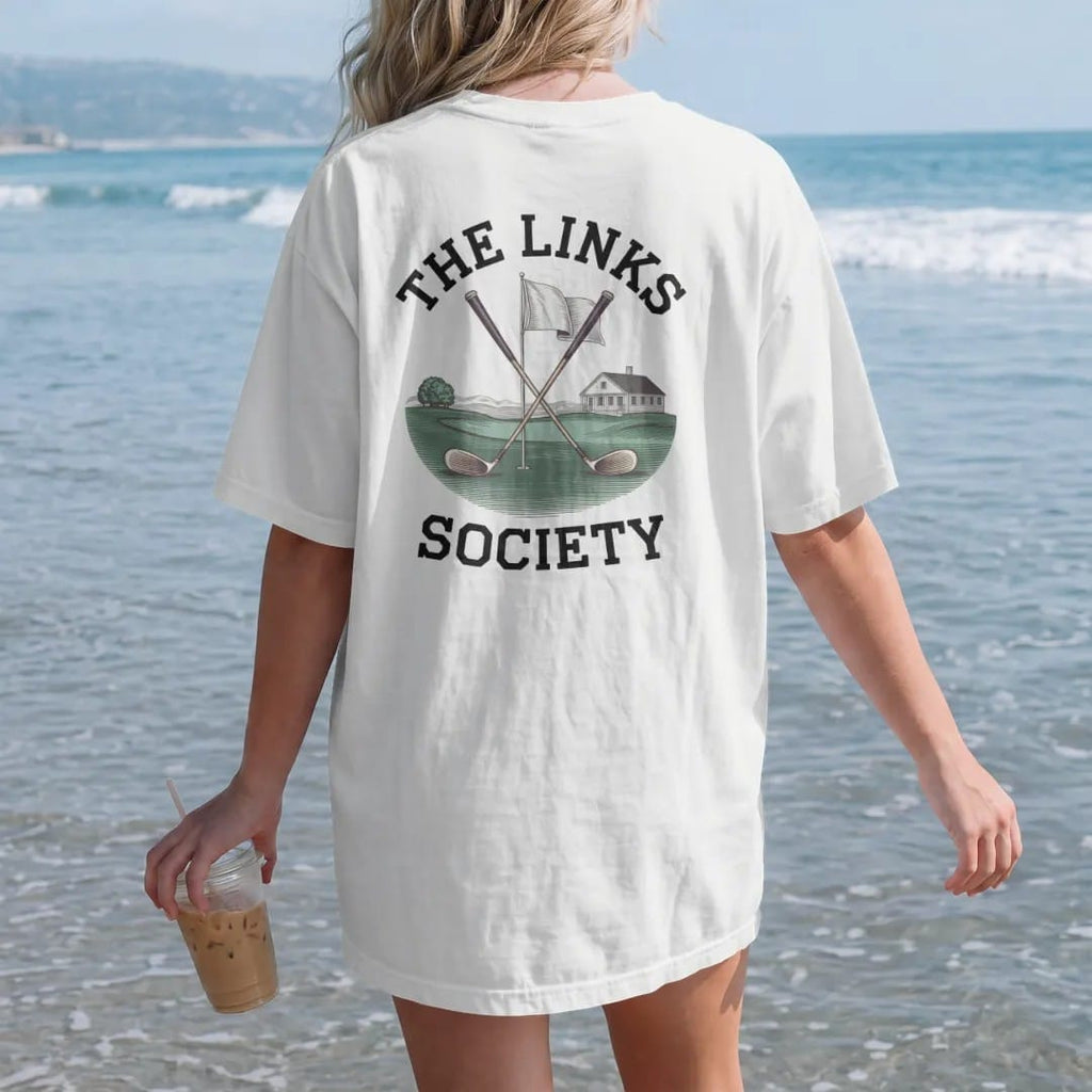 Woman on the beach in a White tee, back view: crossed vintage golf clubs over a flag and green with a clubhouse, under The Links and Society, as a fine engraving in navy and antique gold.