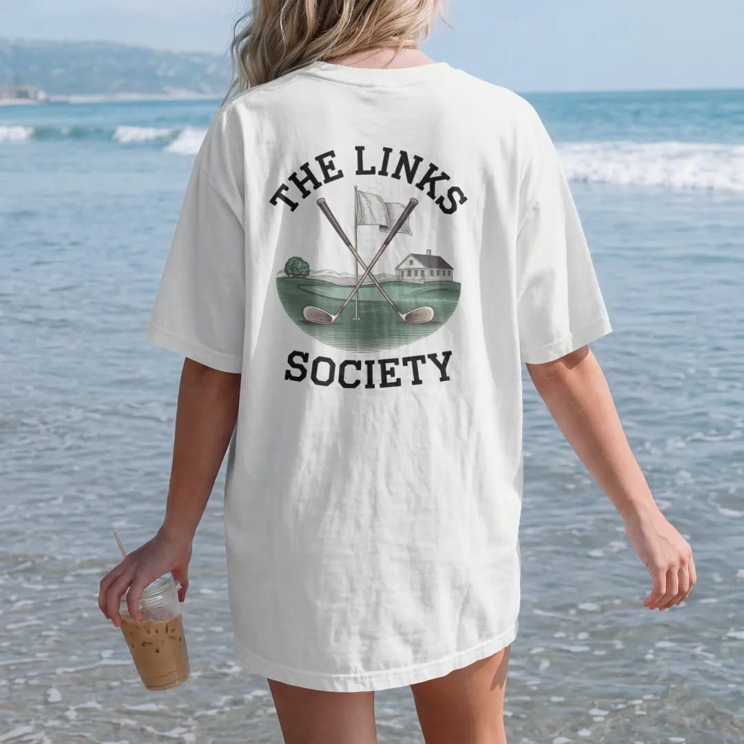 Woman on the beach in a White tee, back view: crossed vintage golf clubs over a flag and green with a clubhouse, under The Links and Society, as a fine engraving in navy and antique gold.