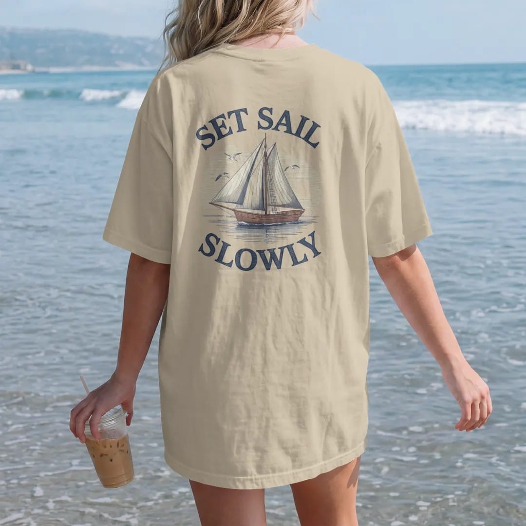 Woman on the beach in a Sandstone tee, back view: a wooden sailboat on calm water with gulls and a low sun, under Set Sail and a script Slowly, as a fine engraving in navy and antique gold.
