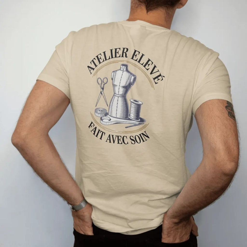 Man wearing a Sandstone tee, seen from the back: a dress form, scissors, tape measure and thread spool under Atelier Elevé and the script Fait Avec Soin, as a fine engraving in navy and antique gold.
