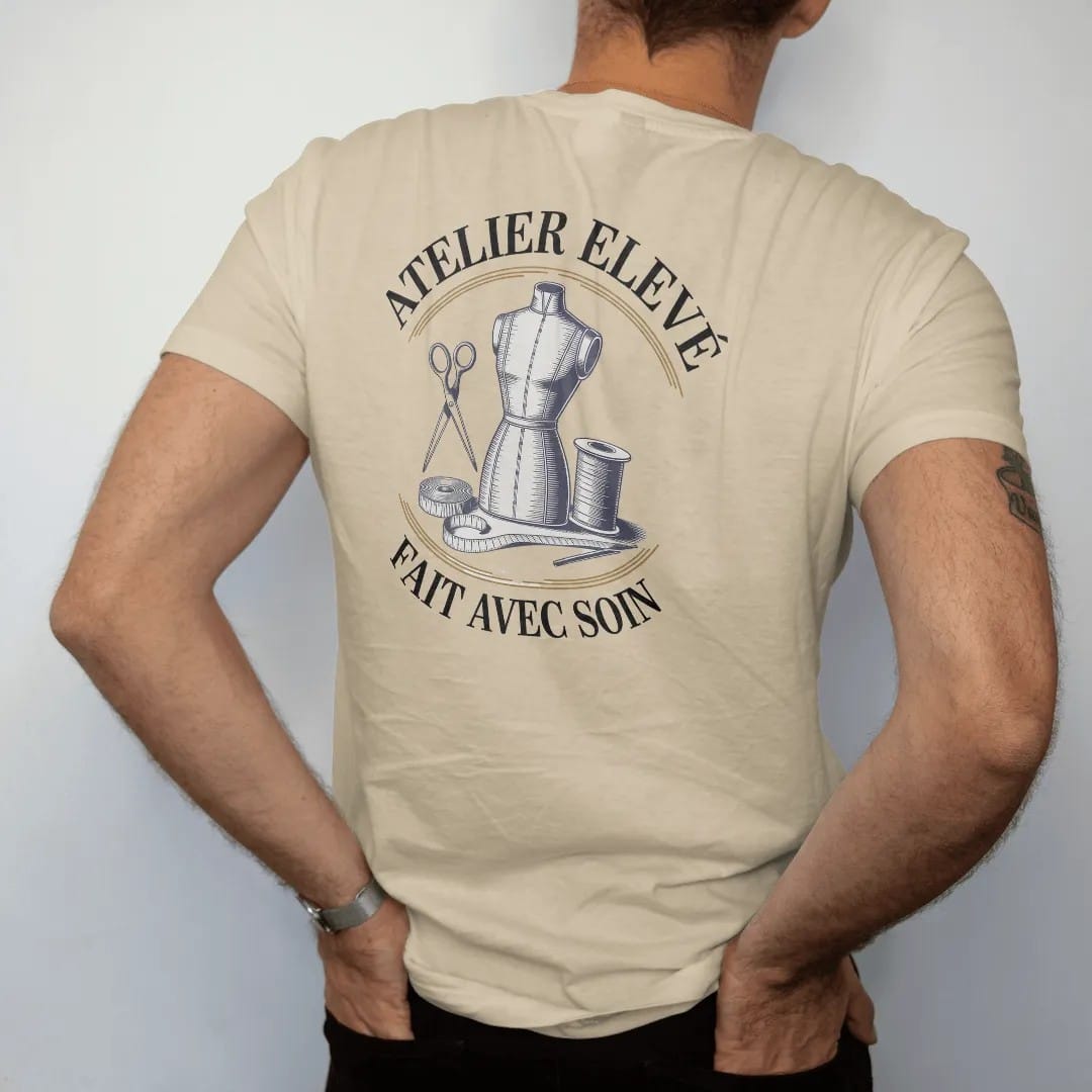 Man wearing a Sandstone tee, seen from the back: a dress form, scissors, tape measure and thread spool under Atelier Elevé and the script Fait Avec Soin, as a fine engraving in navy and antique gold.