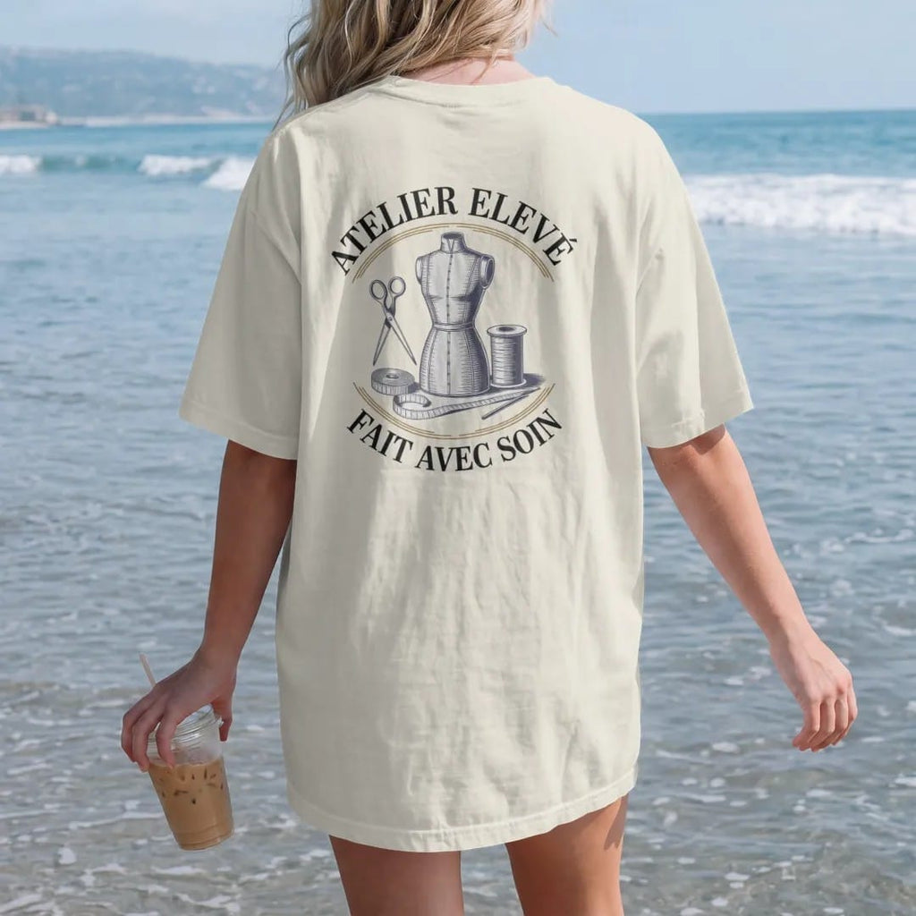 Woman on the beach in a Ivory tee, back view: a dress form, scissors, tape measure and thread spool under Atelier Elevé and the script Fait Avec Soin, as a fine engraving in navy and antique gold.