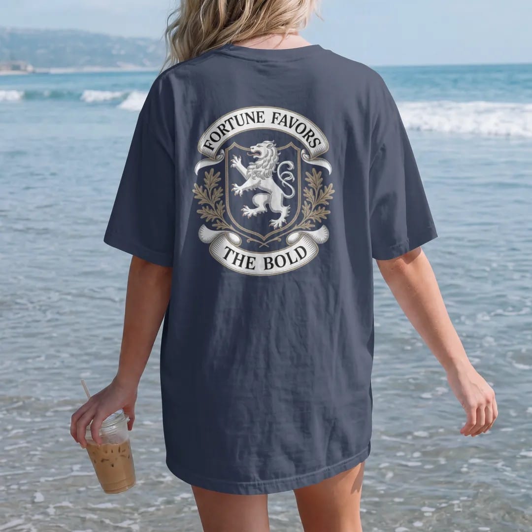 Woman on the beach in a Navy tee, back view: a rampant lion on a shield with oak leaves, Fortune Favors arched above and The Bold on a ribbon, as a fine engraving in cream and champagne gold.