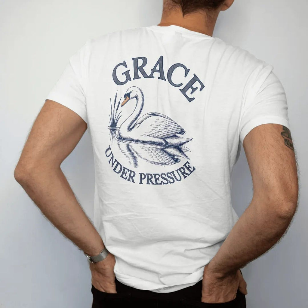 Man wearing a White tee, seen from the back: a swan gliding on still water with reeds and a reflection under Grace in script and Under Pressure, as a fine engraving in navy and antique gold.