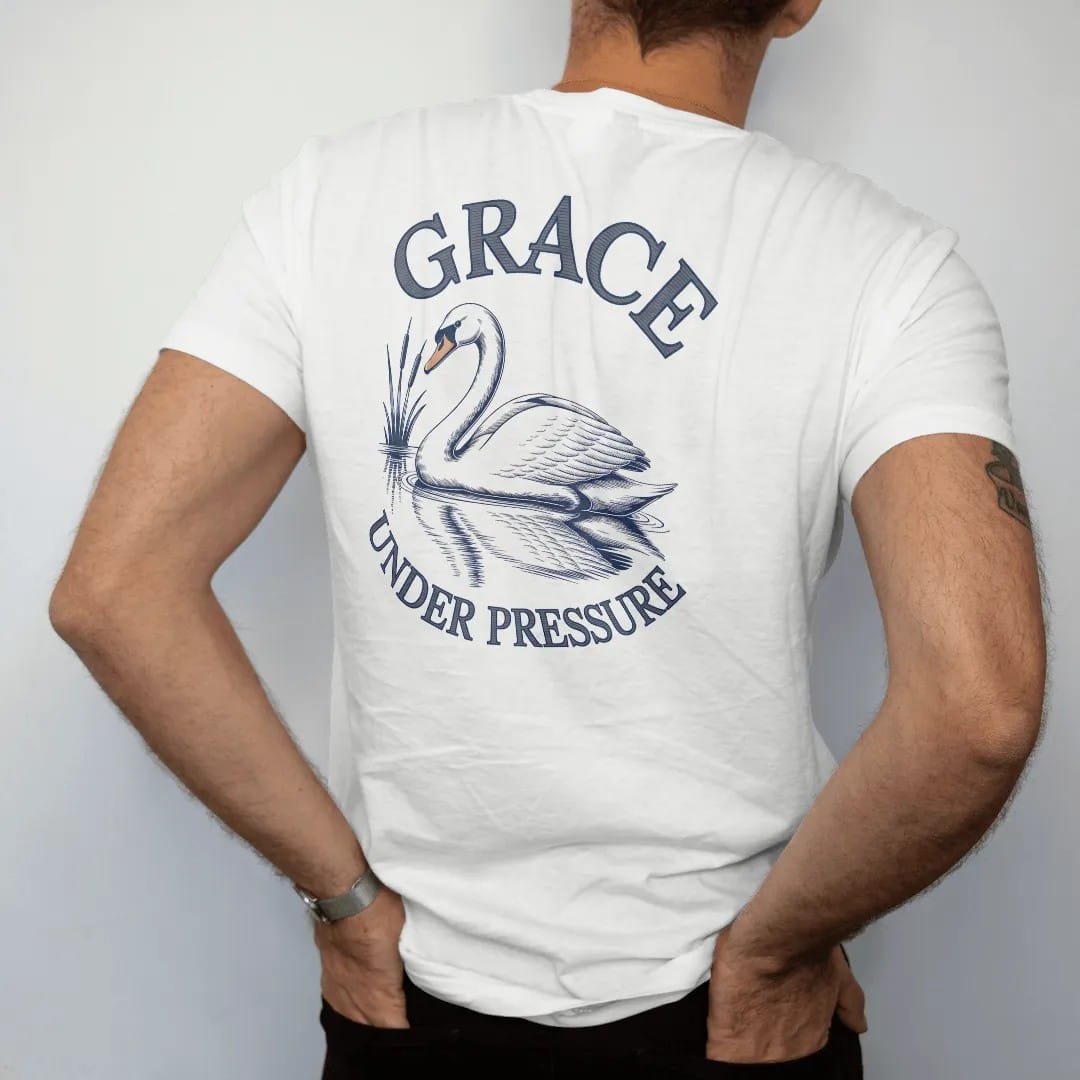 Man wearing a White tee, seen from the back: a swan gliding on still water with reeds and a reflection under Grace in script and Under Pressure, as a fine engraving in navy and antique gold.