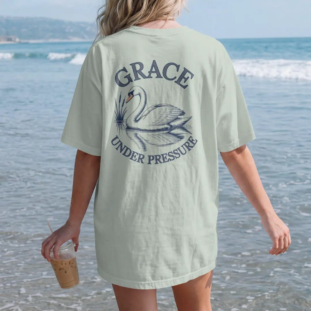 Woman on the beach in a Bay tee, back view: a swan gliding on still water with reeds and a reflection under Grace in script and Under Pressure, as a fine engraving in navy and antique gold.