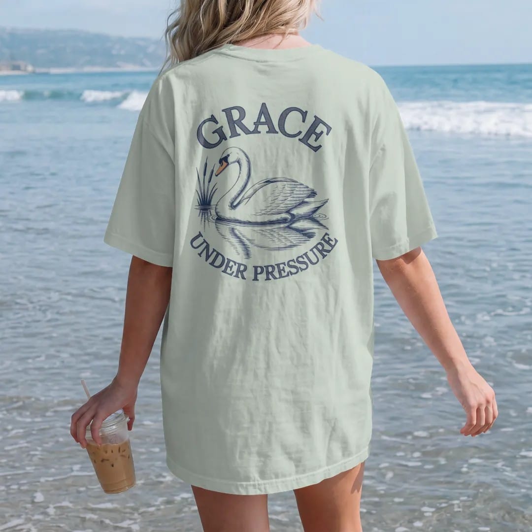 Woman on the beach in a Bay tee, back view: a swan gliding on still water with reeds and a reflection under Grace in script and Under Pressure, as a fine engraving in navy and antique gold.