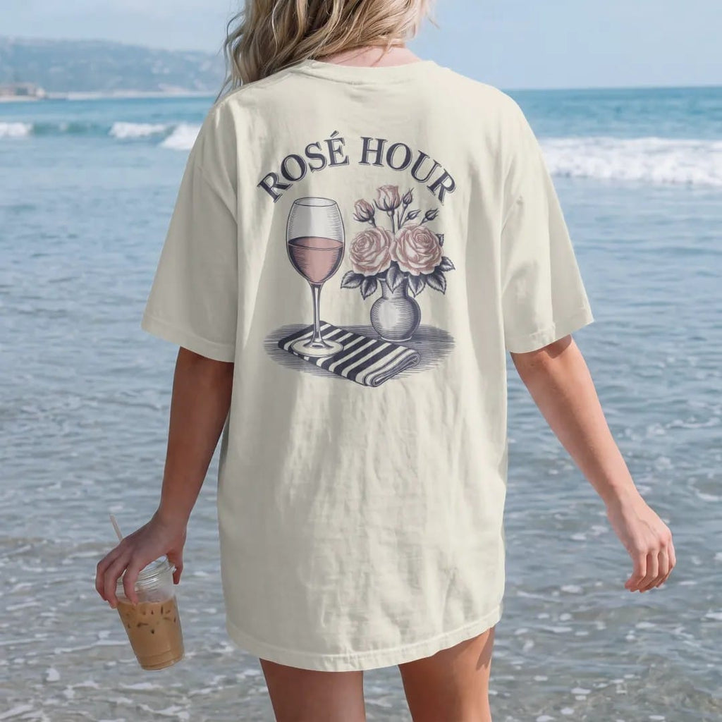 Woman on the beach in a Ivory tee, back view: a glass of rosé beside a vase of garden roses and a striped napkin under a script Rosé Hour, as a fine engraving in navy and antique gold.