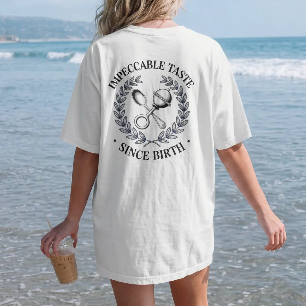 Woman on the beach in a White tee, back view: a silver baby spoon and pearl rattle crossed in a laurel wreath under Impeccable Taste and the script Since Birth, as a fine engraving in navy and antique gold.