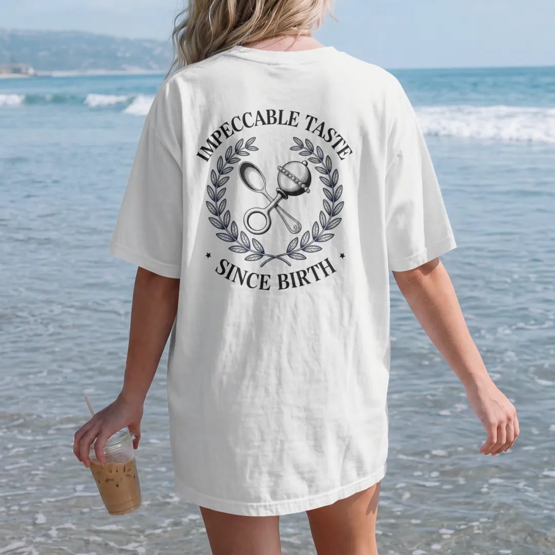 Woman on the beach in a White tee, back view: a silver baby spoon and pearl rattle crossed in a laurel wreath under Impeccable Taste and the script Since Birth, as a fine engraving in navy and antique gold.
