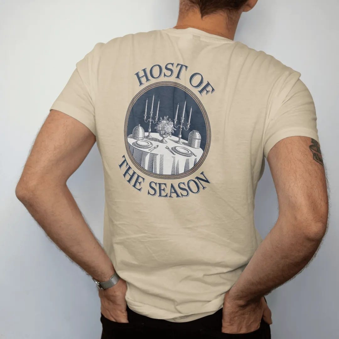 Man wearing a Sandstone tee, seen from the back: a dinner table with candlesticks, a silver cloche and flowers in a round medallion, under Host Of and The Season, as a fine engraving in navy and antique gold.