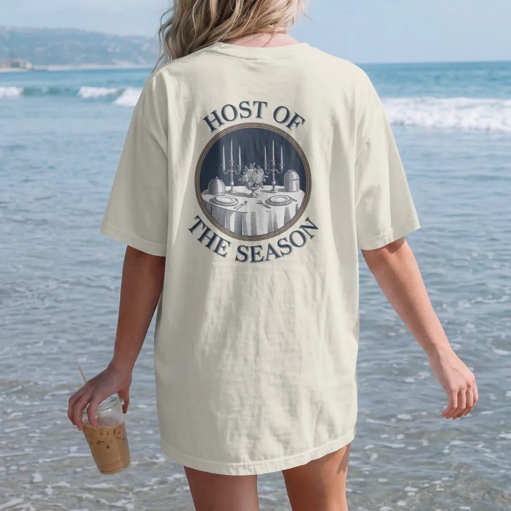 Woman on the beach in a Ivory tee, back view: a dinner table with candlesticks, a silver cloche and flowers in a round medallion, under Host Of and The Season, as a fine engraving in navy and antique gold.