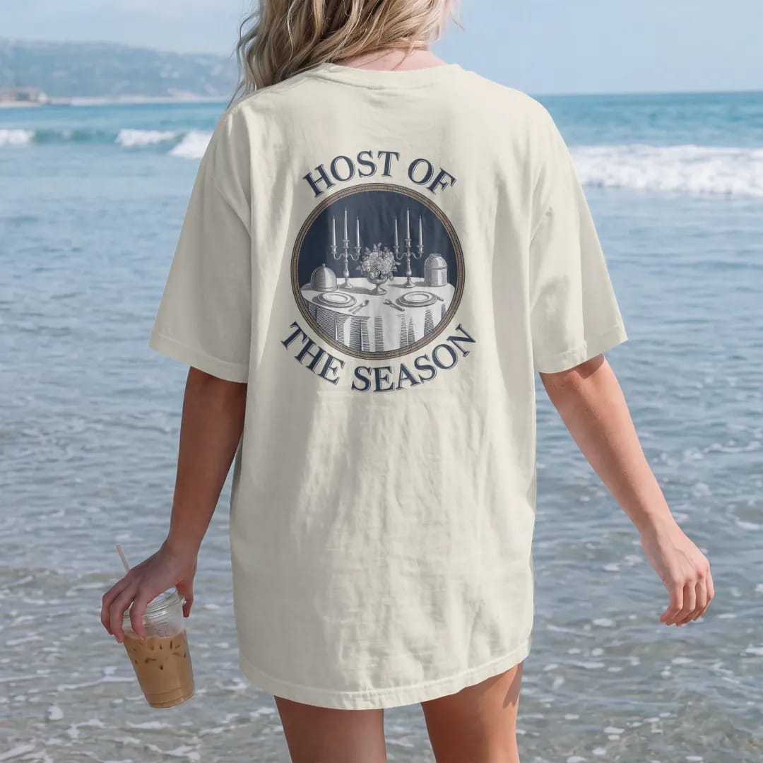Woman on the beach in a Ivory tee, back view: a dinner table with candlesticks, a silver cloche and flowers in a round medallion, under Host Of and The Season, as a fine engraving in navy and antique gold.