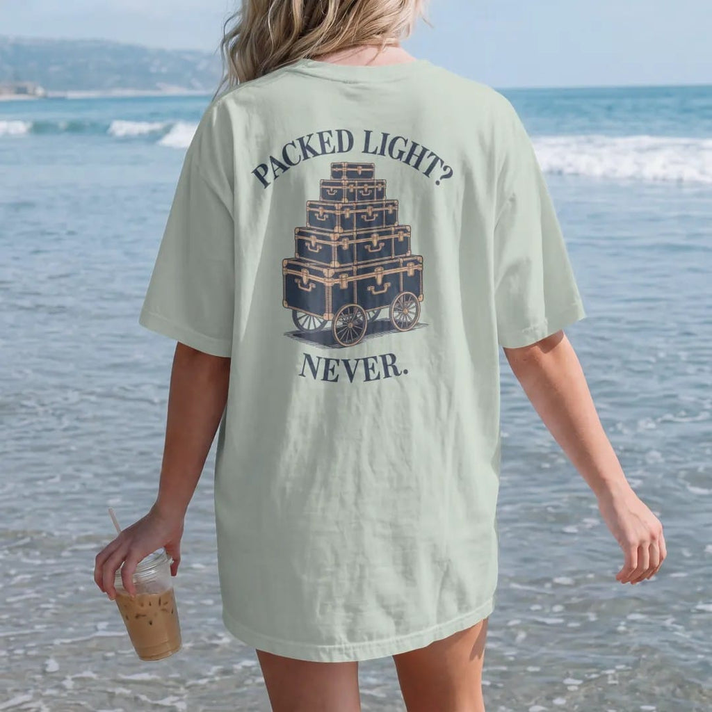 Woman on the beach in a Bay tee, back view: stacked monogrammed steamer trunks and hatboxes on a luggage cart under Packed Light? and Never., as a fine engraving in navy and antique gold.