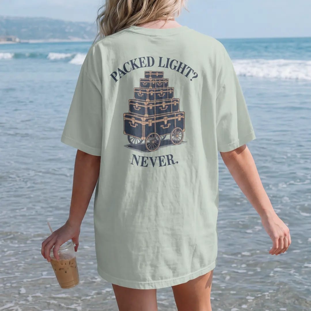 Woman on the beach in a Bay tee, back view: stacked monogrammed steamer trunks and hatboxes on a luggage cart under Packed Light? and Never., as a fine engraving in navy and antique gold.
