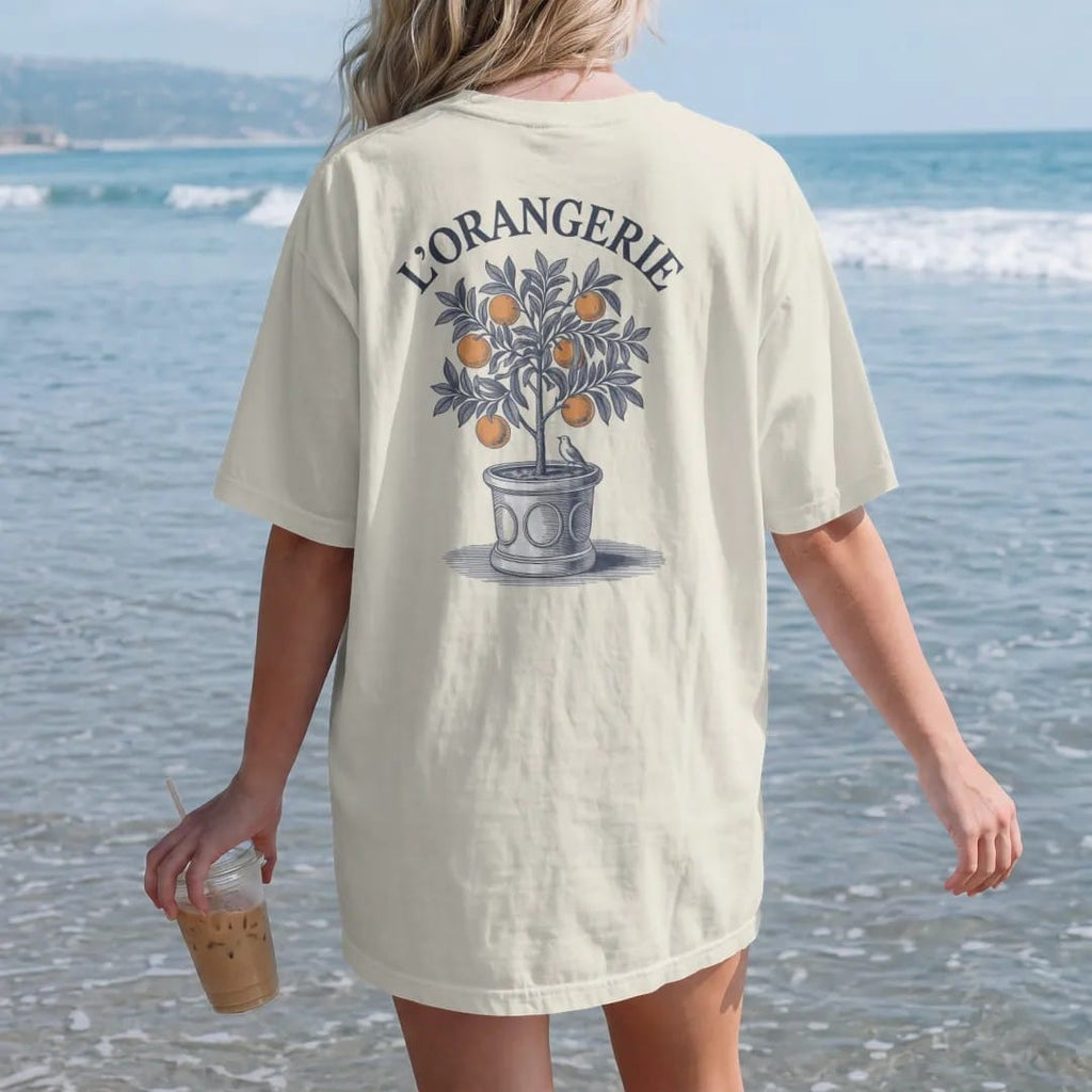 Woman on the beach in a Ivory tee, back view: a potted orange tree with ripe fruit and a small bird in a classic planter box under an arched L'Orangerie, as a fine engraving in navy and antique gold.