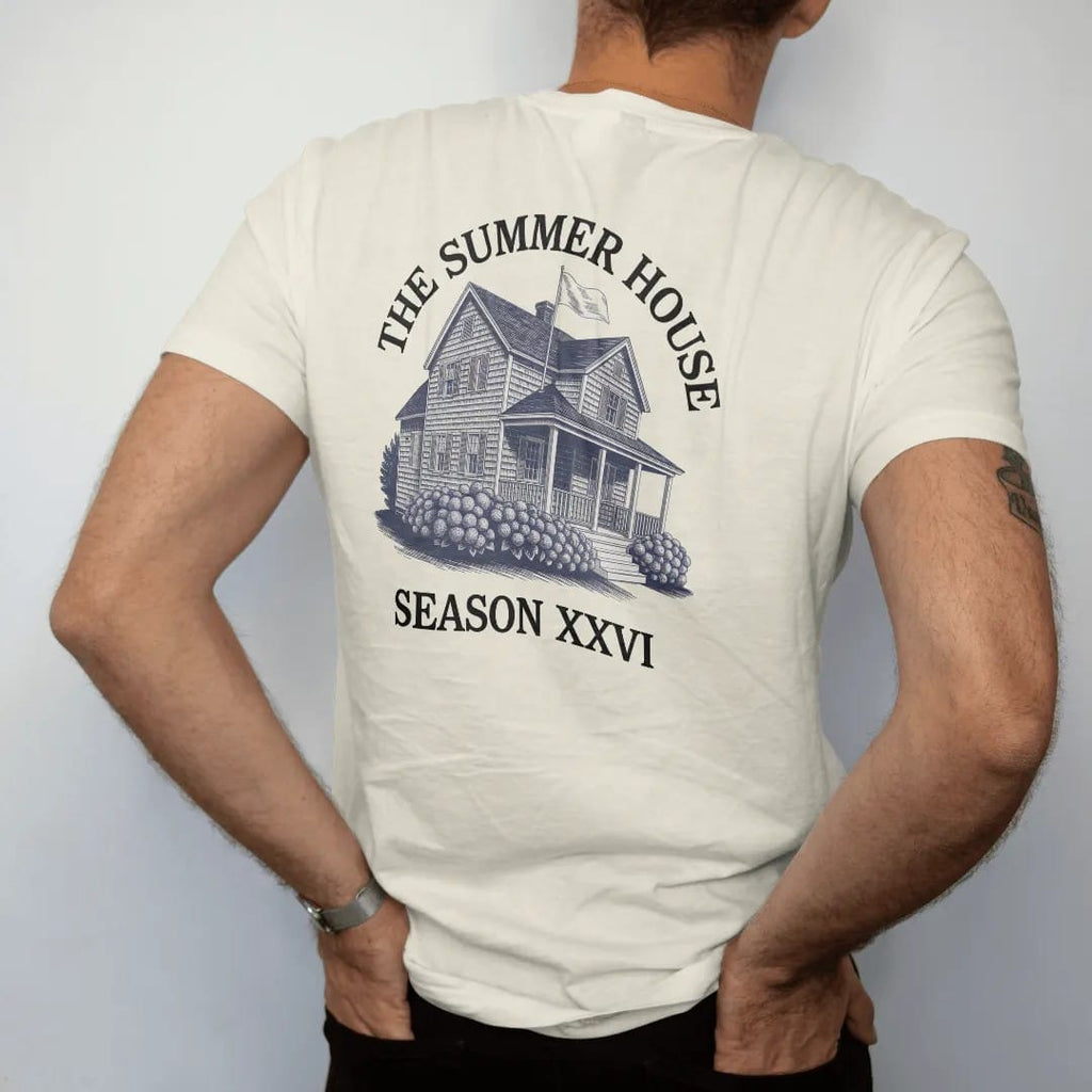 Man wearing a Ivory tee, seen from the back: a shingled summer house with a porch and hydrangeas, under The Summer House and Season XXVI, as a fine engraving in navy and antique gold.